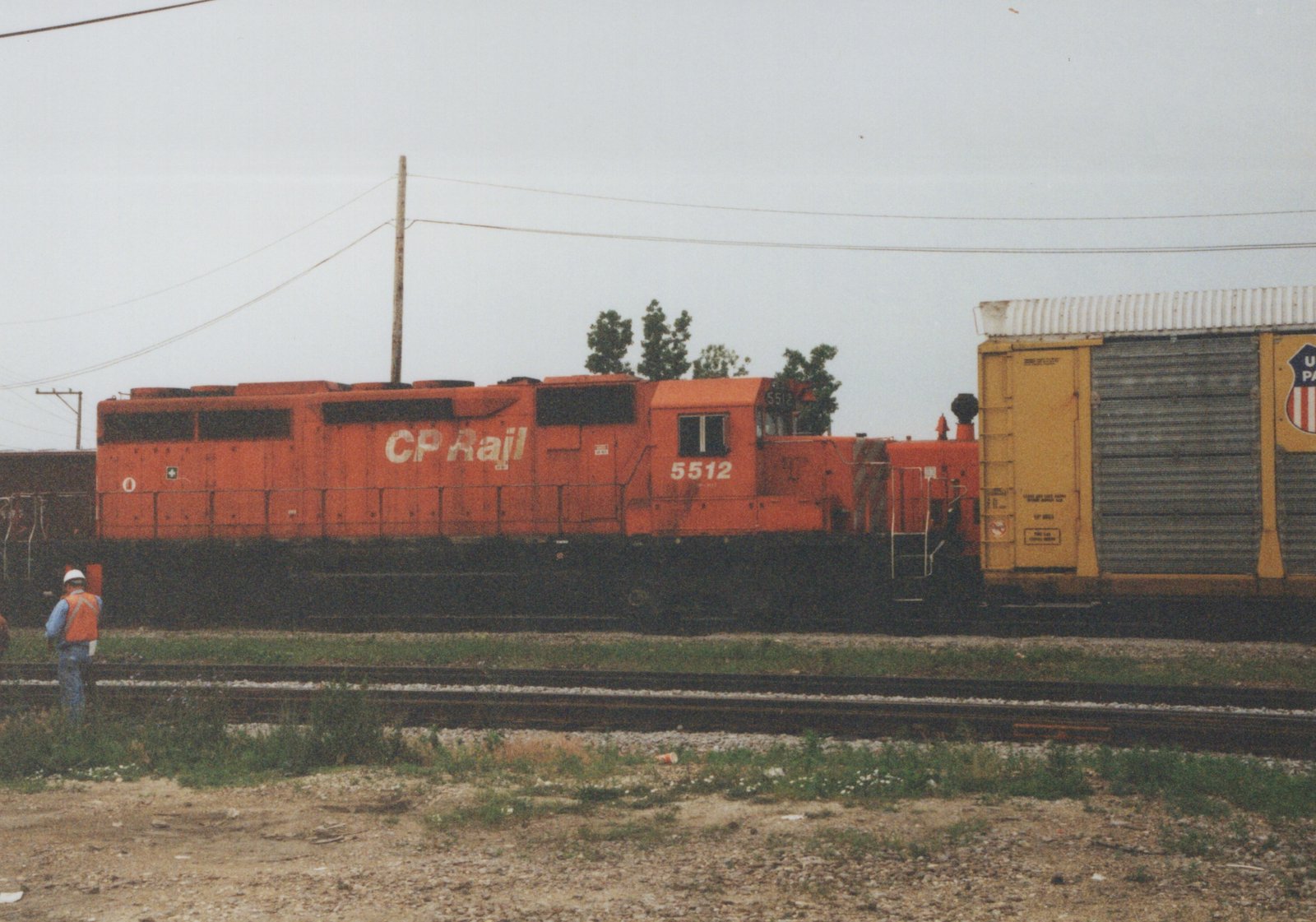 Side view of a red CP Rail diesel locomotive numbered 5512, next to a yellow Union Pacific boxcar, with a person standing near the tracks on an overcast day.