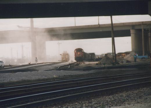 A red and white locomotive pulls a freight train under a large concrete bridge on a dusty, overcast day. Several people and pieces of equipment are visible on the tracks and surrounding area.