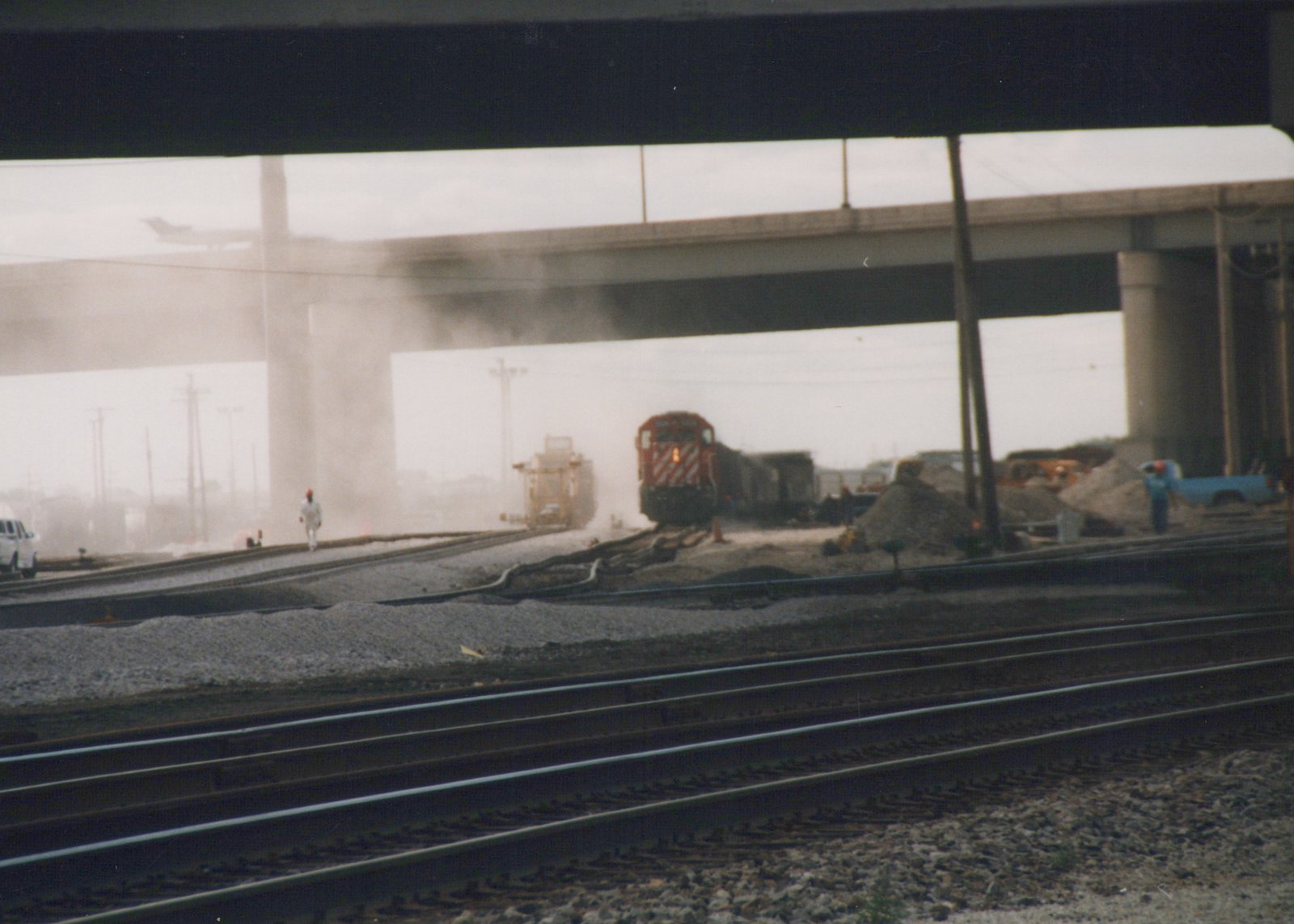 A red and white locomotive pulls a freight train under a large concrete bridge on a dusty, overcast day. Several people and pieces of equipment are visible on the tracks and surrounding area.