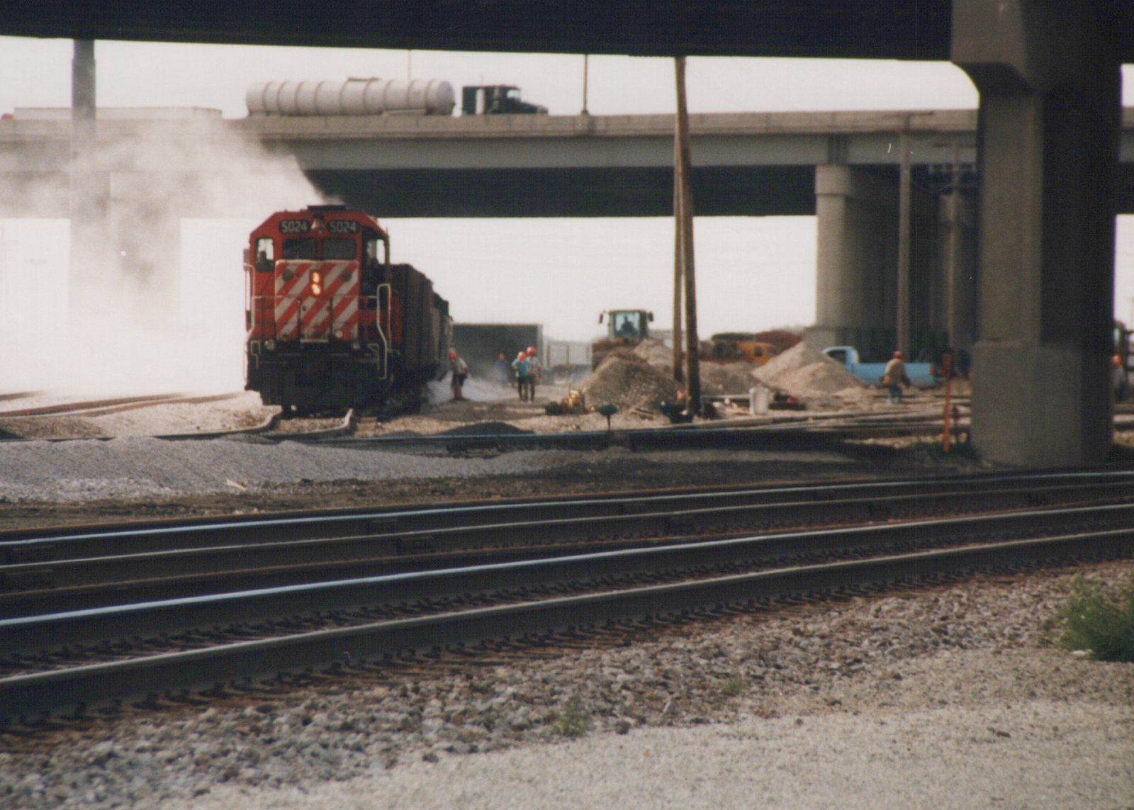 A red diesel locomotive with white diagonal stripes, numbered 5024, is seen moving on tracks under a concrete highway overpass with construction workers and equipment nearby.