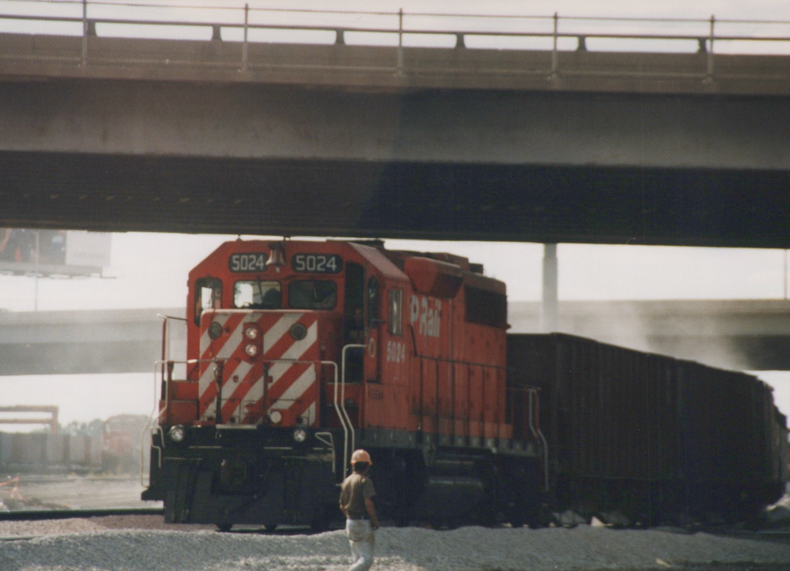 A red diesel locomotive with the number 5024 and 'P Rail' lettering is pulling a line of dark freight cars under a concrete bridge, with one person standing on the gravel beside the tracks.