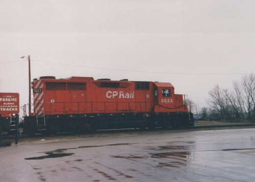 Side view of a red CP Rail locomotive numbered 5023, parked on a wet pavement next to railroad tracks, with bare trees and a 'No Parking' sign visible.