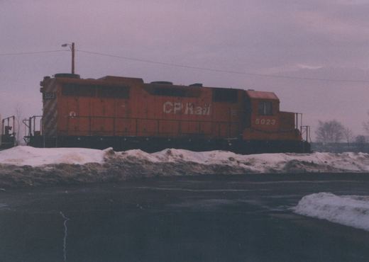 A stationary orange Canadian Pacific diesel locomotive numbered 5023, seen from the side, with snow piled beside the tracks under a gray sky.