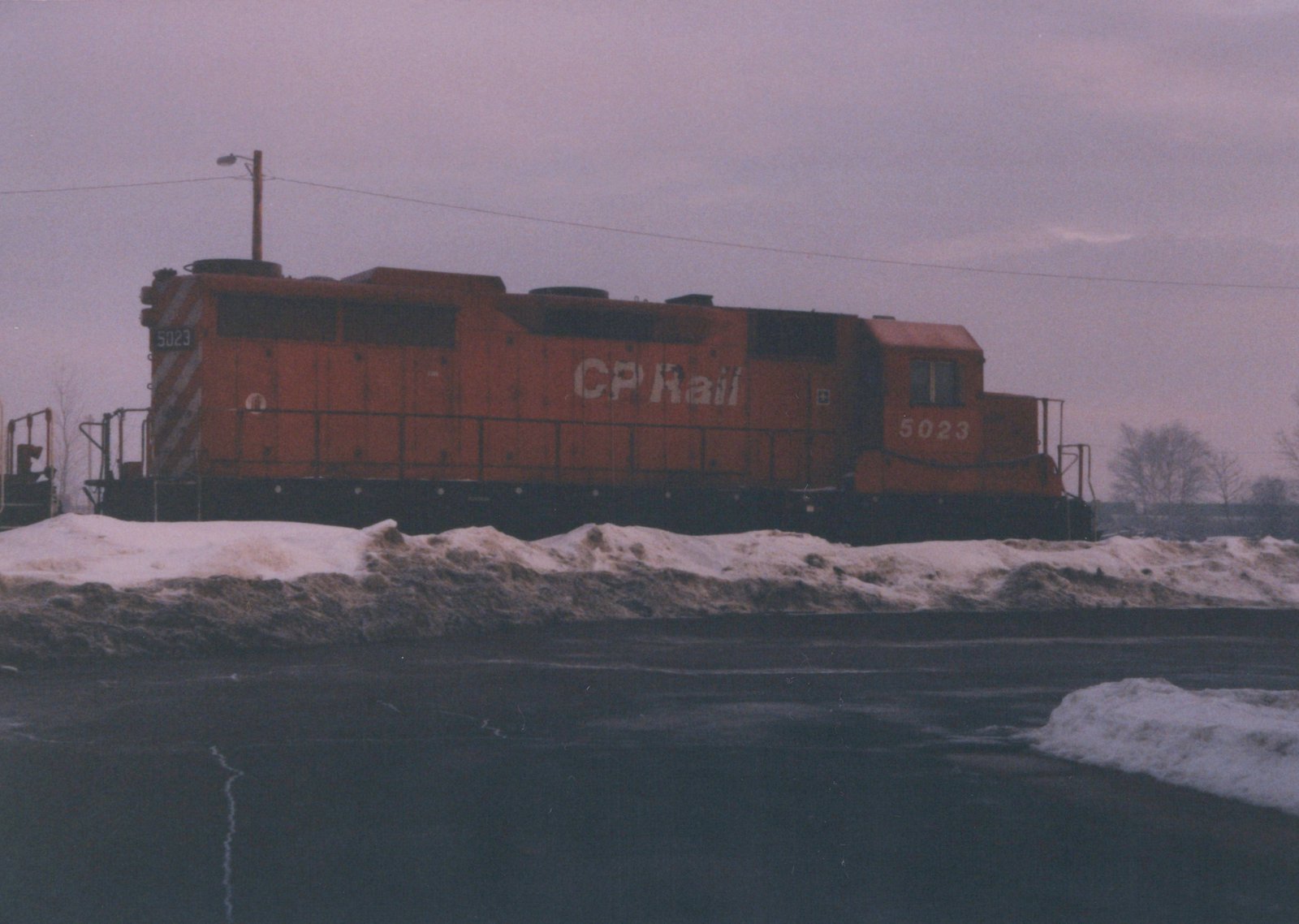 A stationary orange Canadian Pacific diesel locomotive numbered 5023, seen from the side, with snow piled beside the tracks under a gray sky.