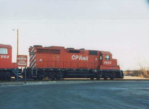 A side view of a red diesel locomotive with 'CP Rail' written on its side, numbered 5023, stopped on railroad tracks next to a road sign that reads 'NO PARKING ALONG RAILROAD TRACKS'. A red caboose numbered 202 is partially visible behind it.
