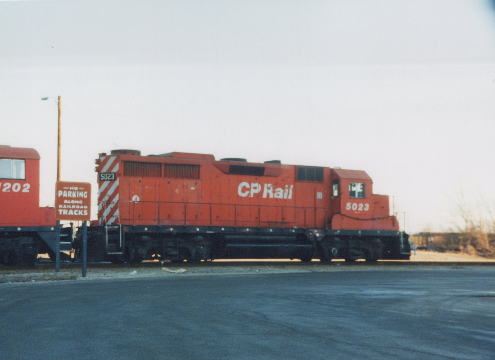 A side view of a red diesel locomotive with 'CP Rail' written on its side, numbered 5023, stopped on railroad tracks next to a road sign that reads 'NO PARKING ALONG RAILROAD TRACKS'. A red caboose numbered 202 is partially visible behind it.