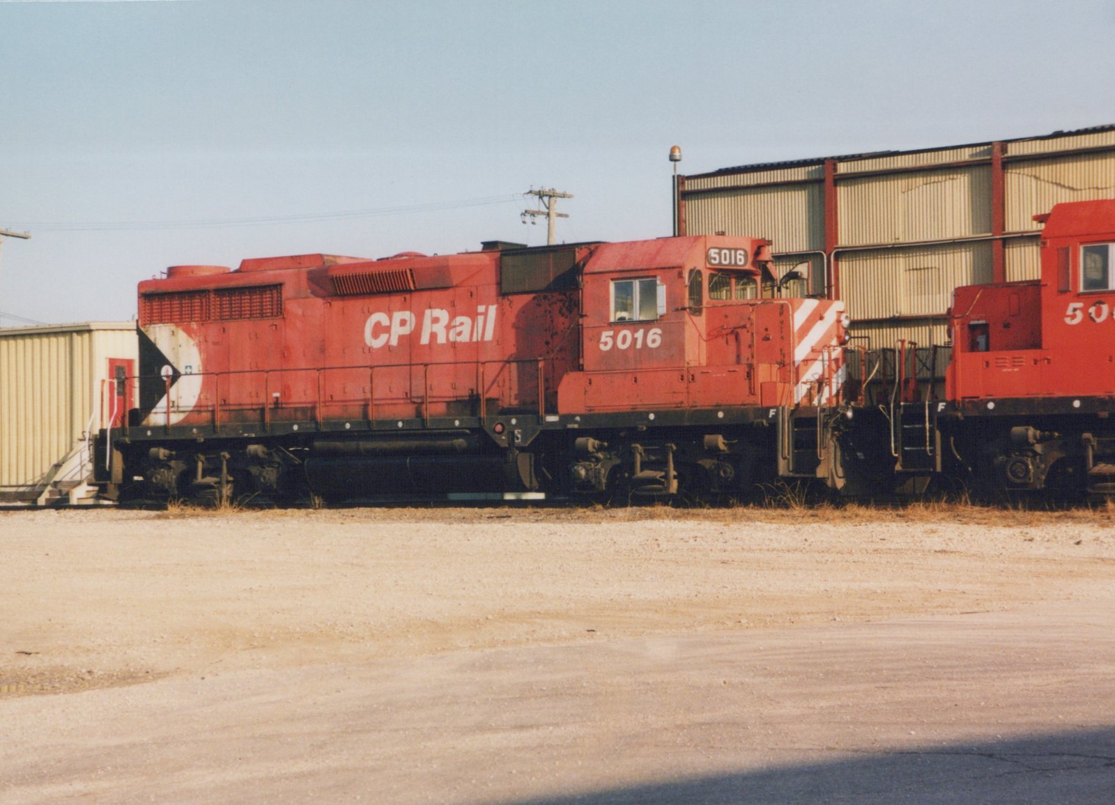 Side view of two red Canadian Pacific diesel locomotives in a gravel yard, with industrial buildings in the background.