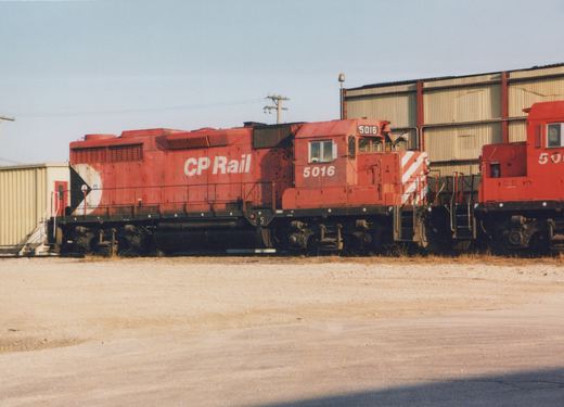 Side view of two red Canadian Pacific diesel locomotives in a gravel yard, with industrial buildings in the background.