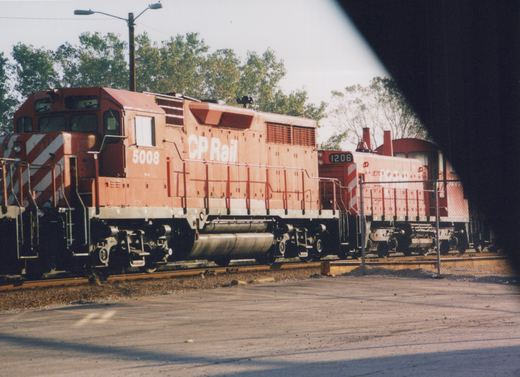Side view of two red Canadian Pacific diesel locomotives, number 5008 in front and 1206 behind, parked on parallel tracks in a sunny yard with trees in the background.