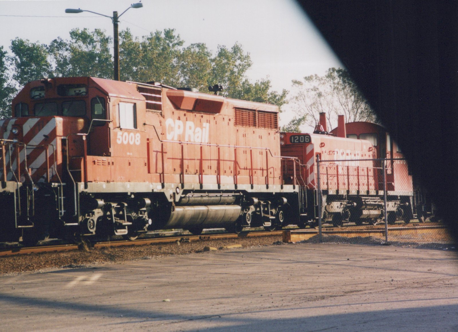 Side view of two red Canadian Pacific diesel locomotives, number 5008 in front and 1206 behind, parked on parallel tracks in a sunny yard with trees in the background.