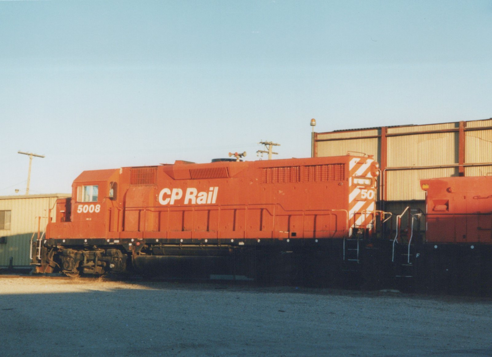 An orange CP Rail diesel locomotive numbered 5008 is parked on a track in a yard next to a building under a clear sky.