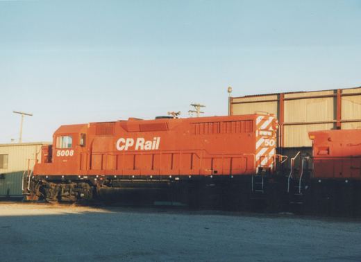 An orange CP Rail diesel locomotive numbered 5008 is parked on a track in a yard next to a building under a clear sky.