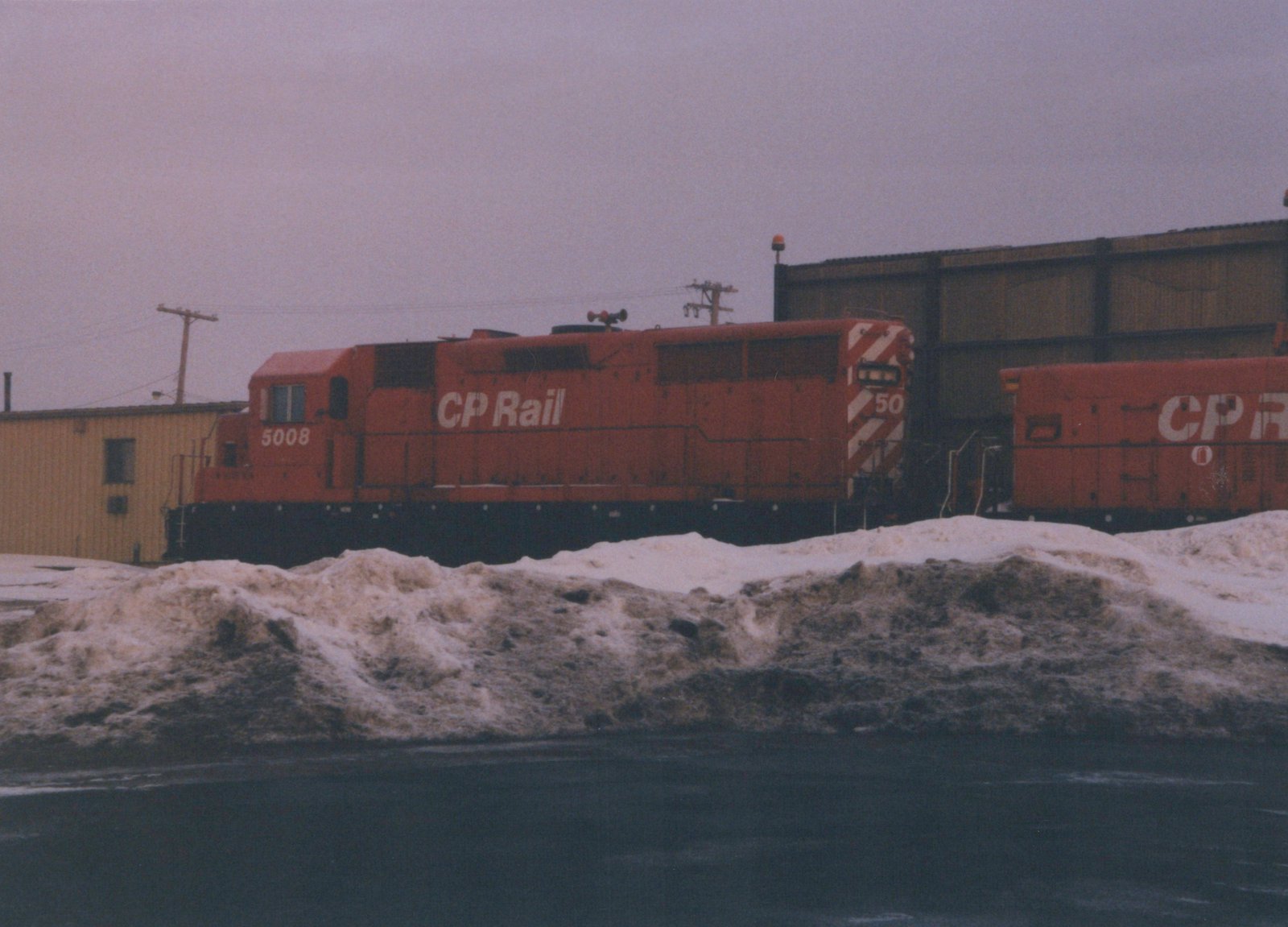 A red Canadian Pacific diesel locomotive numbered 5008 is parked in a snowy yard, with another red CP locomotive partially visible beside it.