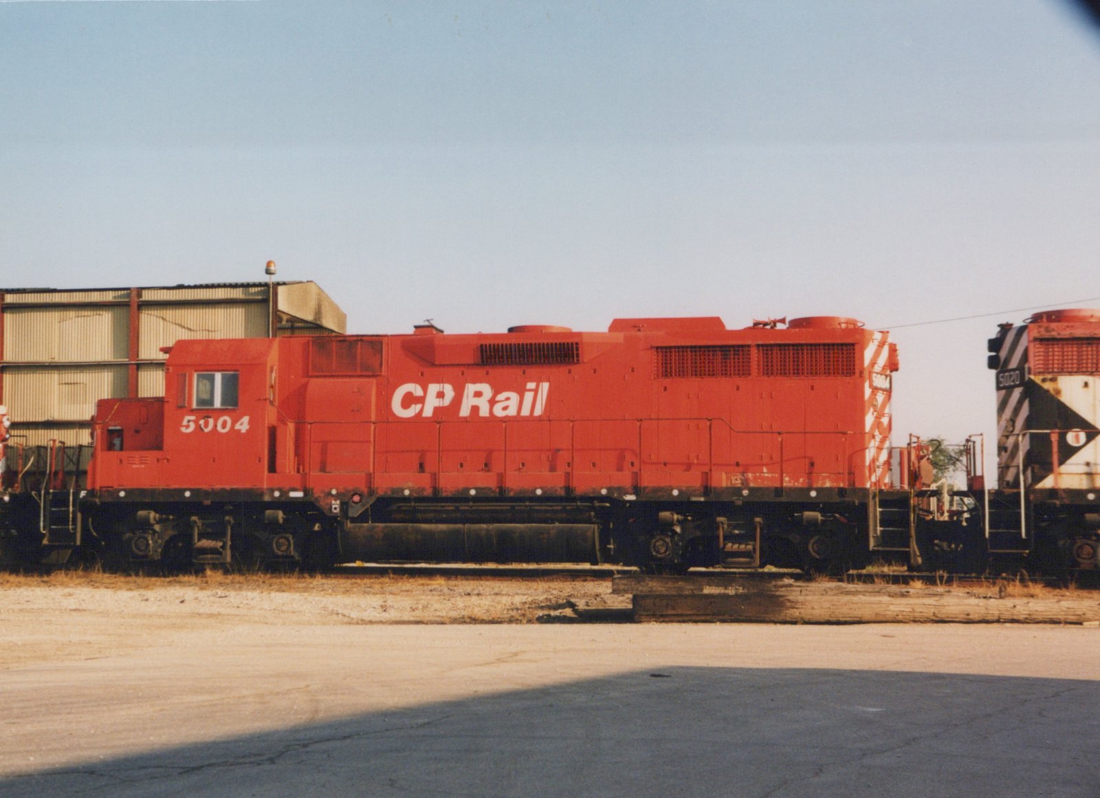 Side view of a bright red Canadian Pacific diesel locomotive numbered 5004, parked in a yard with another locomotive numbered 5020 partially visible in the background.