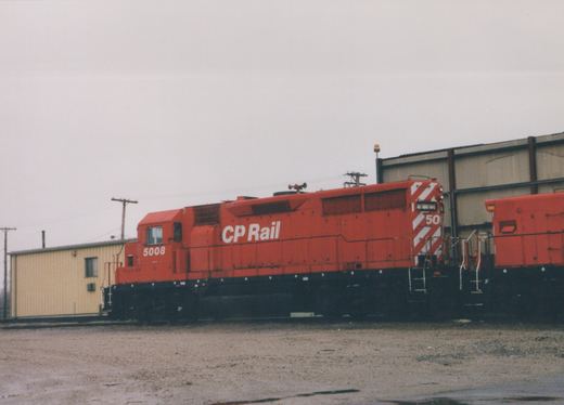 A red Canadian Pacific diesel locomotive with the number 5008 on its side, parked in a gravel yard next to a building, with another locomotive partially visible behind it.