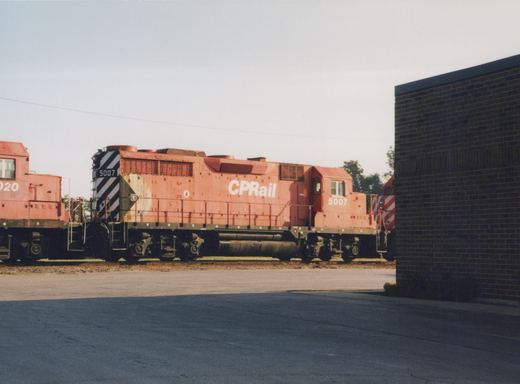 A red Canadian Pacific diesel locomotive numbered 5007 sits on tracks beside a brick building in a yard, viewed from the side.
