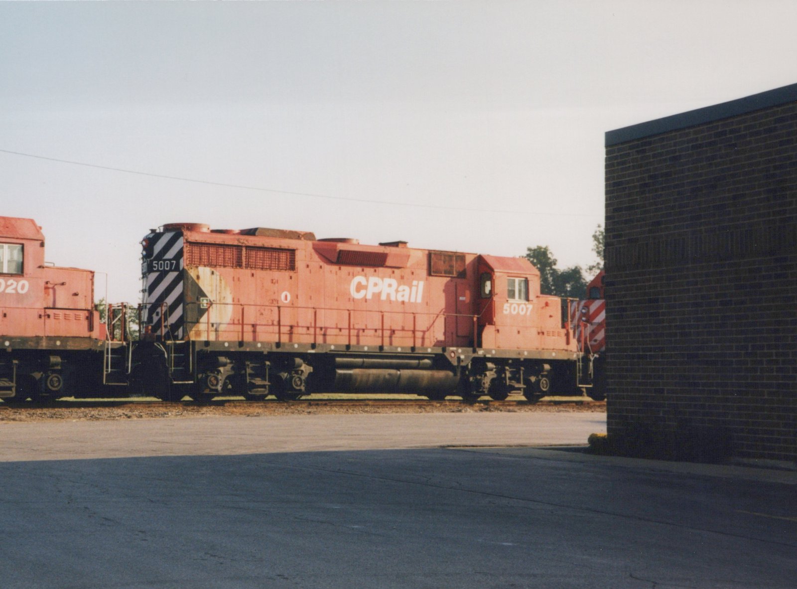 A red Canadian Pacific diesel locomotive numbered 5007 sits on tracks beside a brick building in a yard, viewed from the side.