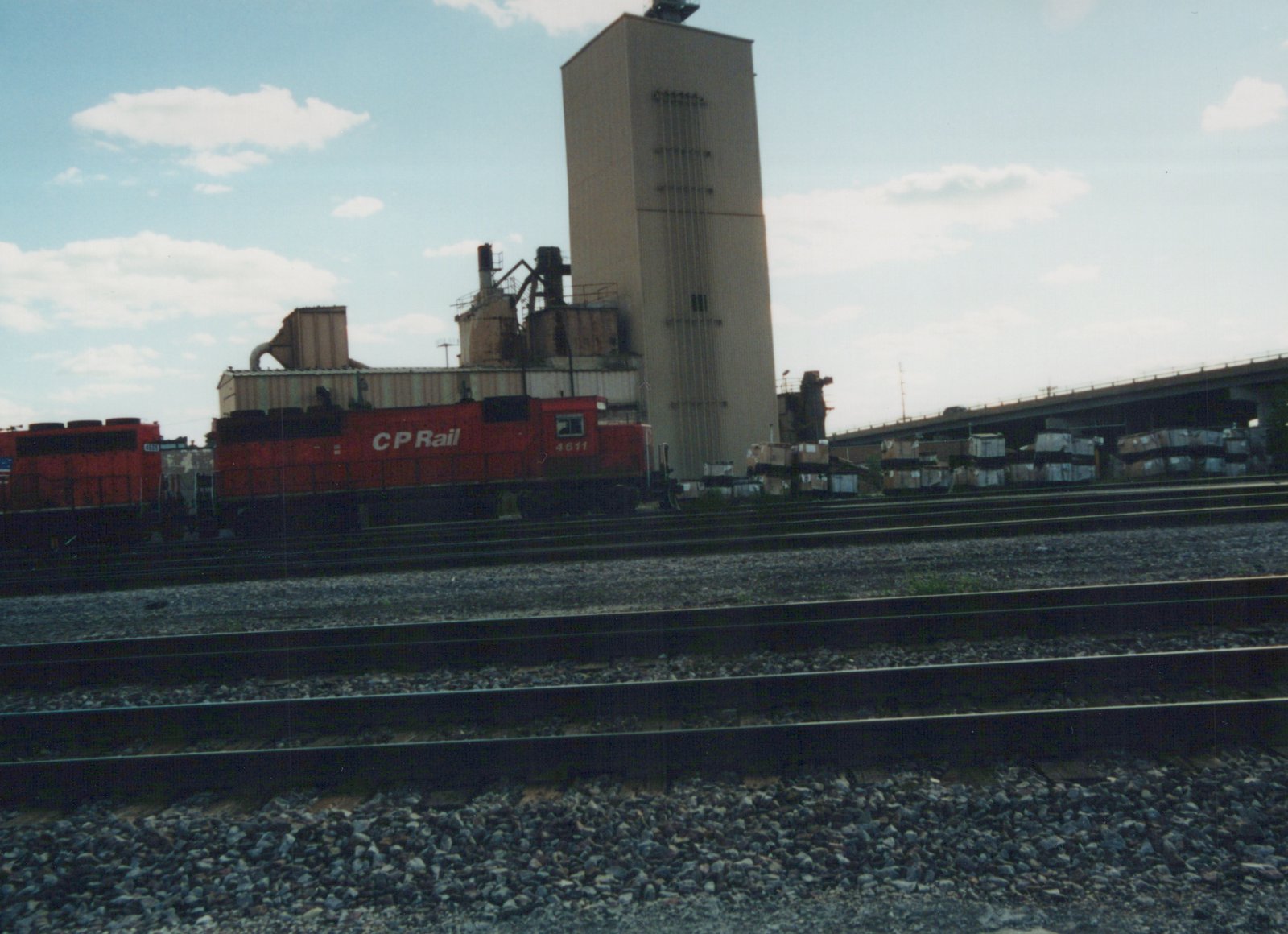 A red Canadian Pacific locomotive numbered 4611 sits on tracks in a yard with gravel ballast, a tall industrial building, and a highway overpass in the background under a partly cloudy sky.