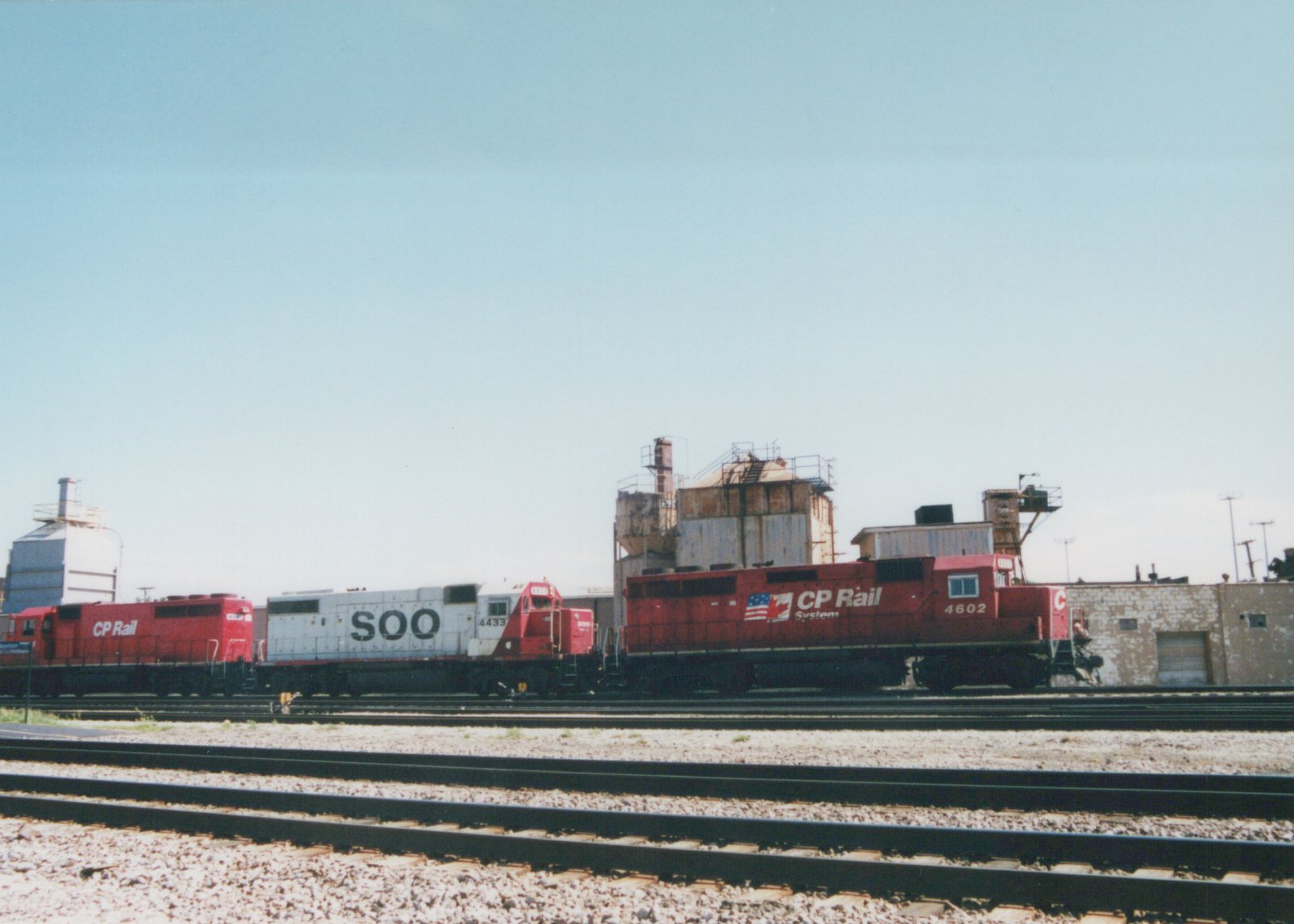 Three diesel locomotives, one white with 'SOO' lettering and two red with 'CP Rail' lettering, are parked in a rail yard on a bright day.