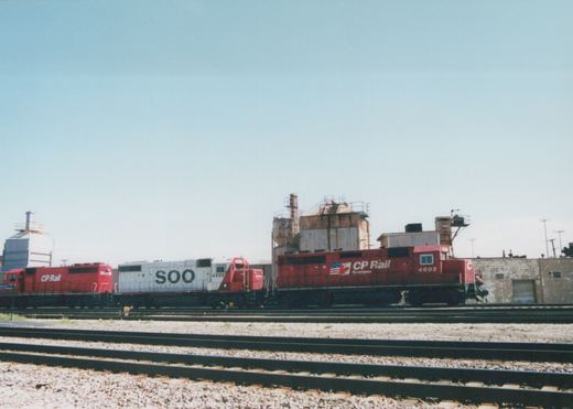 Three diesel locomotives, one white with 'SOO' lettering and two red with 'CP Rail' lettering, are parked in a rail yard on a bright day.