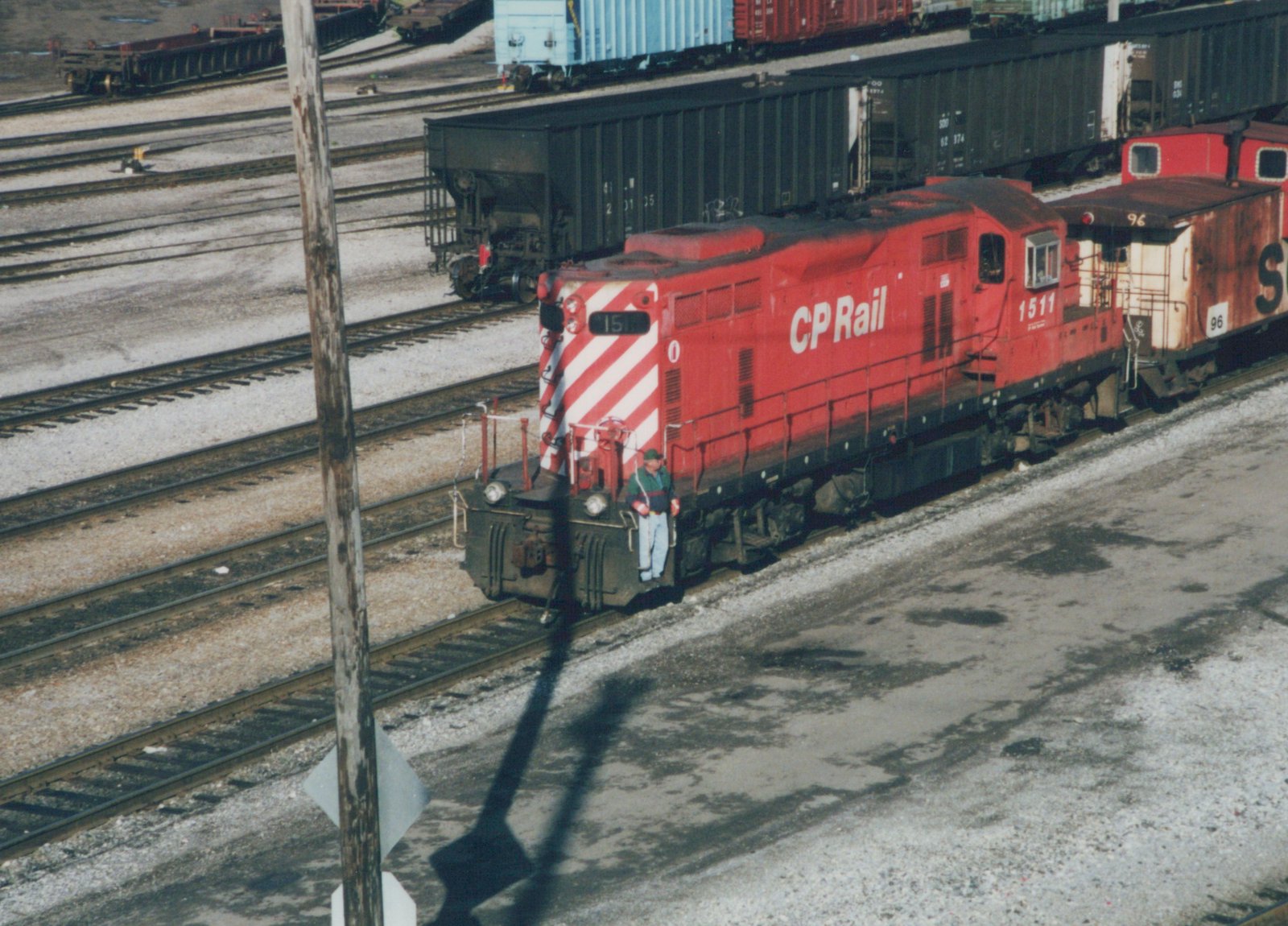 A red CP Rail diesel locomotive, number 1511, is parked in a rail yard with a caboose. A person stands on the front platform. The ground is bare and gravelly, indicating winter.