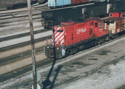 A red CP Rail diesel locomotive, number 1511, is parked in a rail yard with a caboose. A person stands on the front platform. The ground is bare and gravelly, indicating winter.