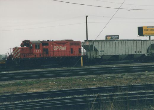 A red Canadian Pacific locomotive numbered 1511 is parked next to a white Great Northern freight car on multiple railway tracks under an overcast sky.
