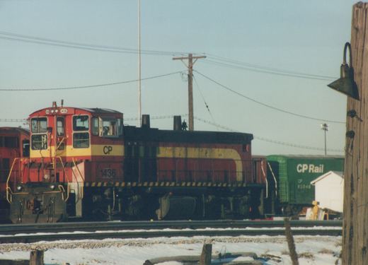 A red and yellow Canadian Pacific diesel locomotive, number 1436, stands on the tracks in a snowy yard next to a green freight car.