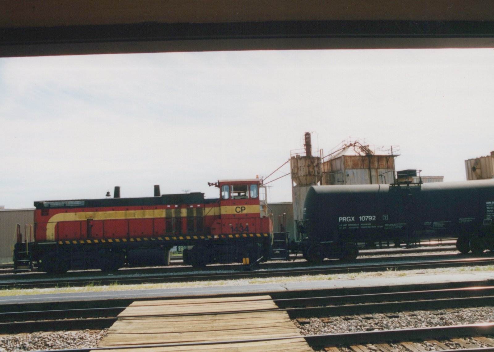 A red and yellow Canadian Pacific locomotive numbered 1434 is parked on railroad tracks next to a black tank car marked PRGX 10792, viewed from under a bridge on an overcast day.