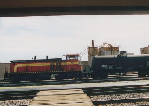 A red and yellow Canadian Pacific locomotive numbered 1434 is parked on railroad tracks next to a black tank car marked PRGX 10792, viewed from under a bridge on an overcast day.