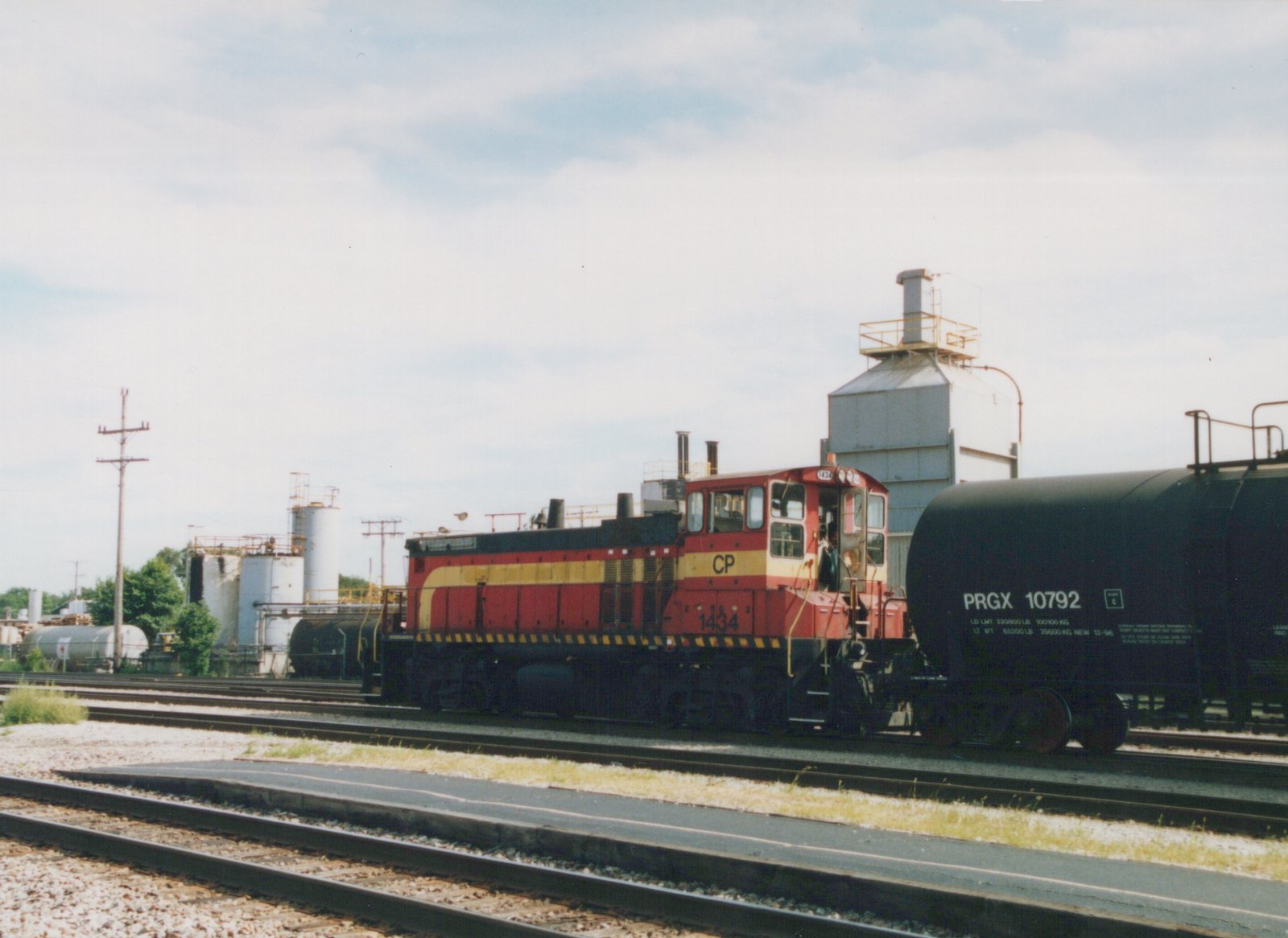 A red and yellow Canadian Pacific locomotive numbered 1434 is coupled to a black tank car in a rail yard with industrial tanks in the background.