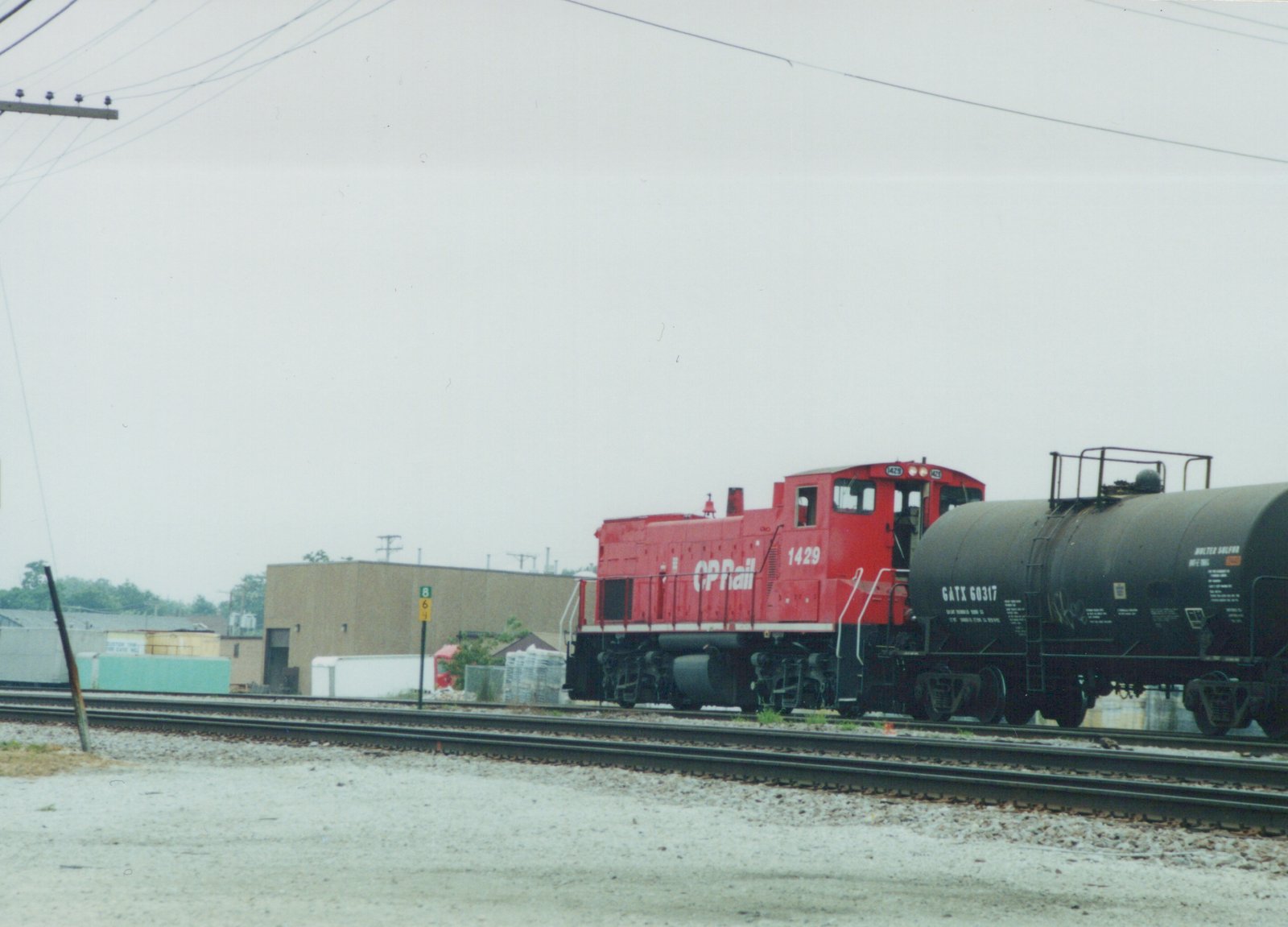A red Canadian Pacific SW1200 locomotive numbered 1429 is pulling a black GATX tank car on a set of railroad tracks in a yard.