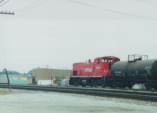 A red Canadian Pacific SW1200 locomotive numbered 1429 is pulling a black GATX tank car on a set of railroad tracks in a yard.