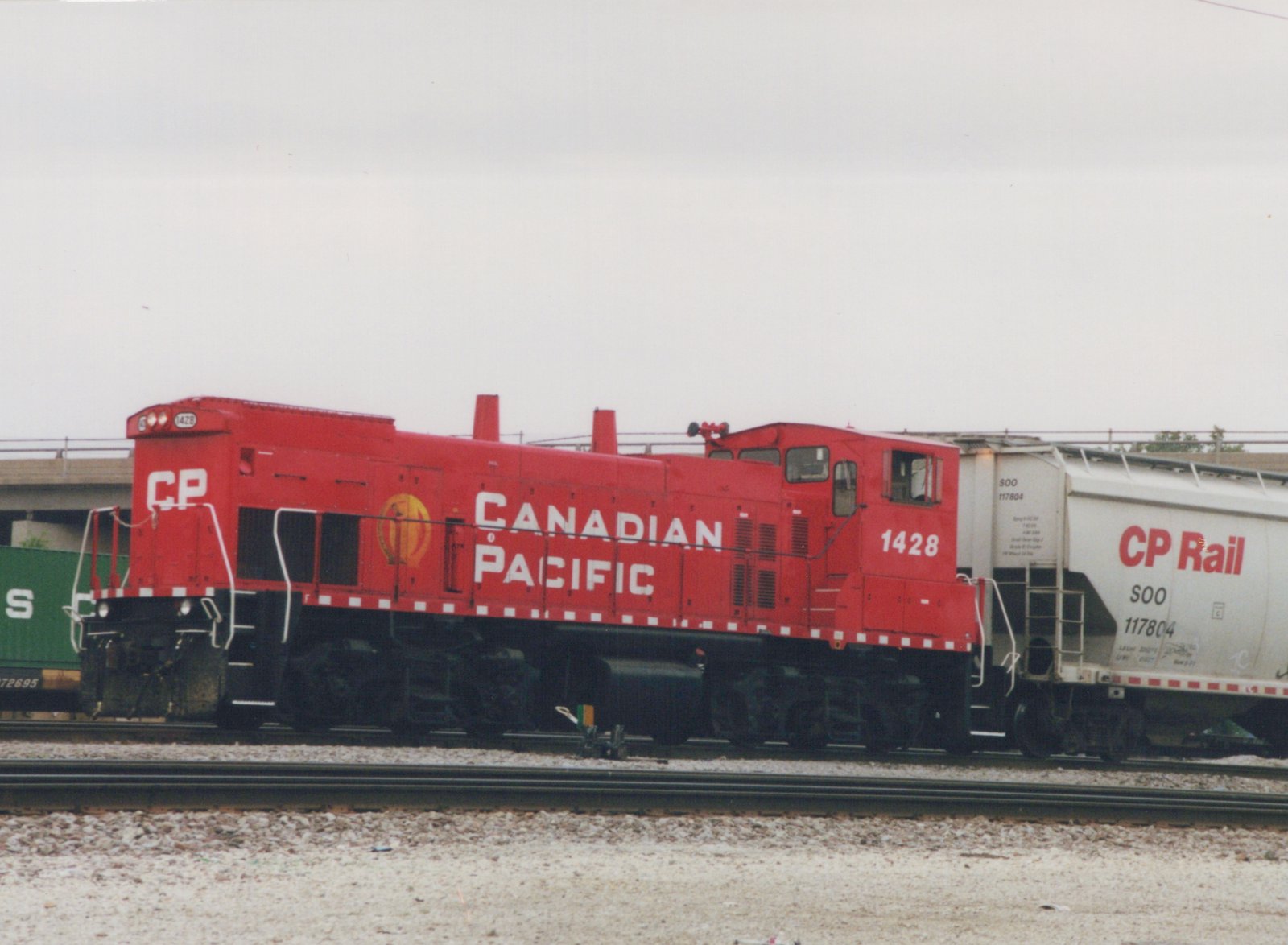 A red Canadian Pacific locomotive number 1428 pulling white and green freight cars on a set of tracks with a concrete overpass in the background under a cloudy sky.