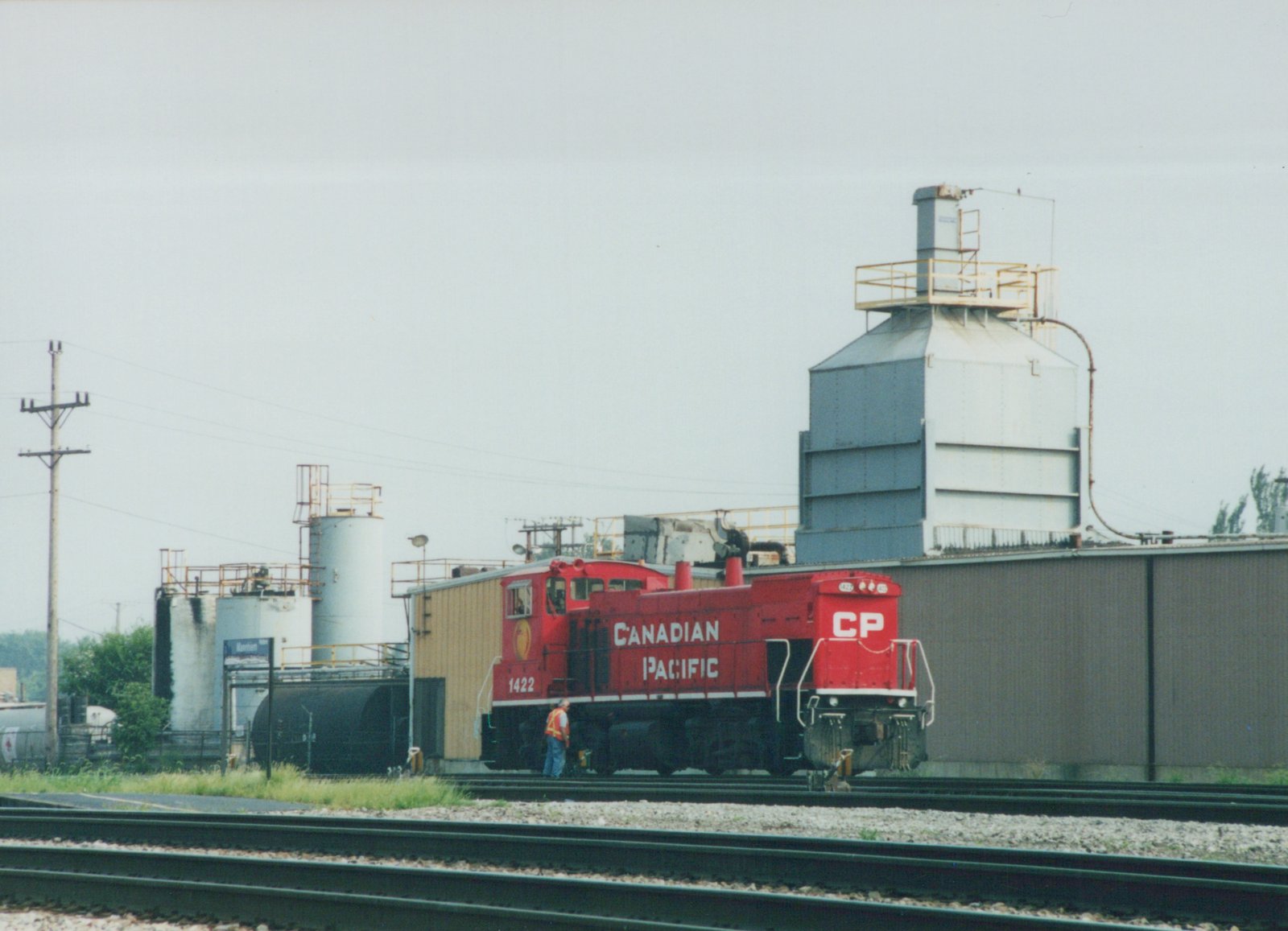 A red Canadian Pacific diesel locomotive number 1422 is parked on railroad tracks next to industrial storage tanks under an overcast sky.