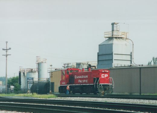 A red Canadian Pacific diesel locomotive number 1422 is parked on railroad tracks next to industrial storage tanks under an overcast sky.