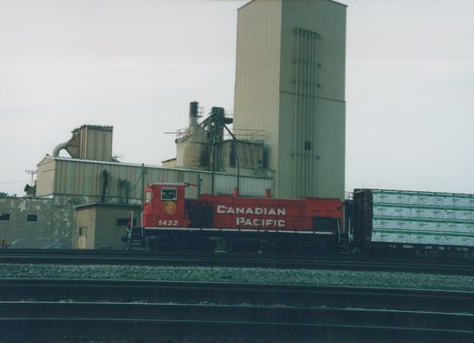 A red Canadian Pacific locomotive numbered 1422 sits on railroad tracks next to a freight car, with industrial buildings in the background.