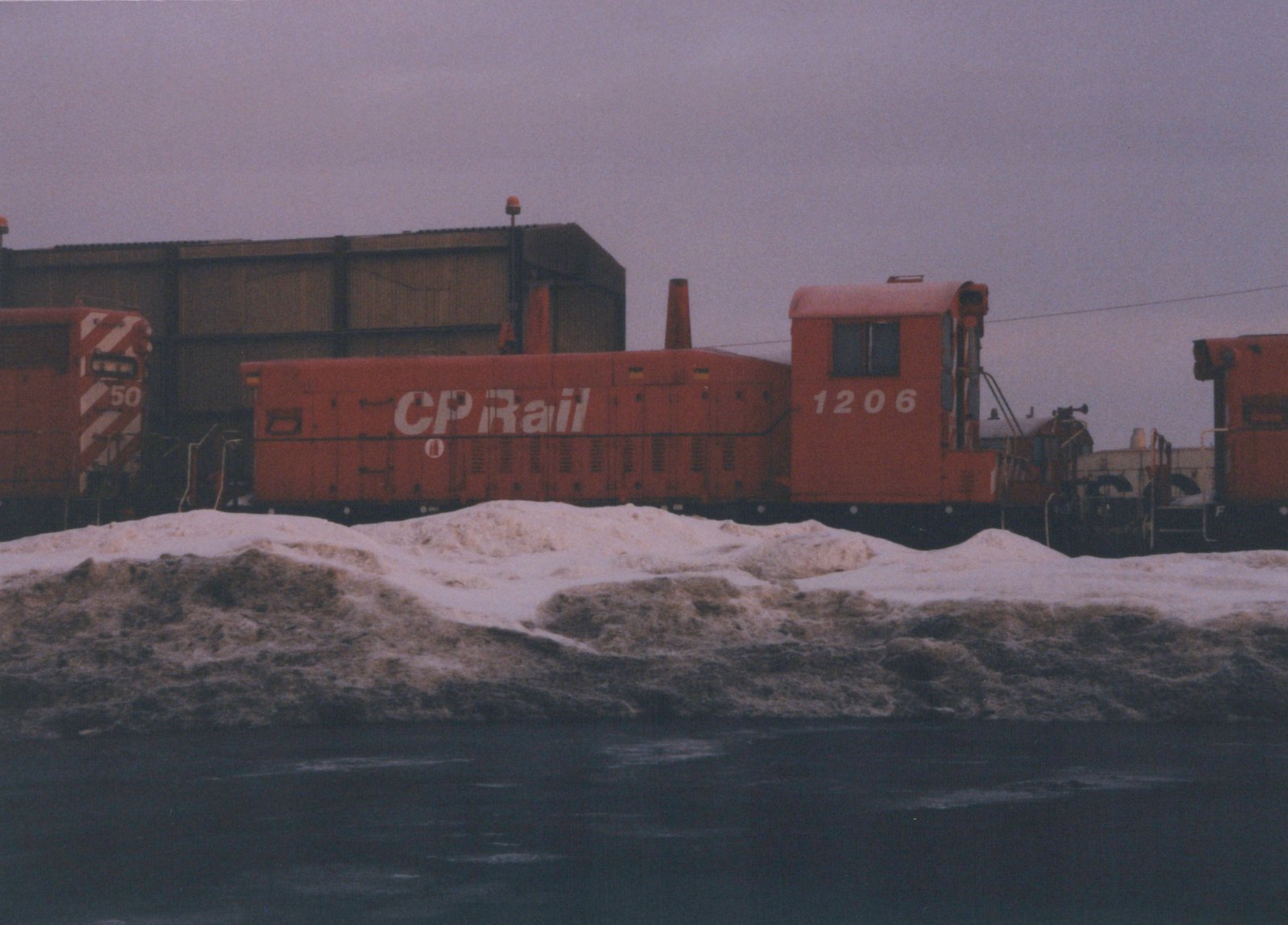 A red diesel locomotive with 'CP Rail' and the number 1206 on its side is parked in a snowy yard, with another locomotive numbered 50 visible nearby.