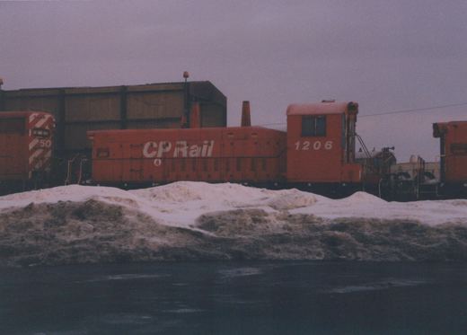 A red diesel locomotive with 'CP Rail' and the number 1206 on its side is parked in a snowy yard, with another locomotive numbered 50 visible nearby.