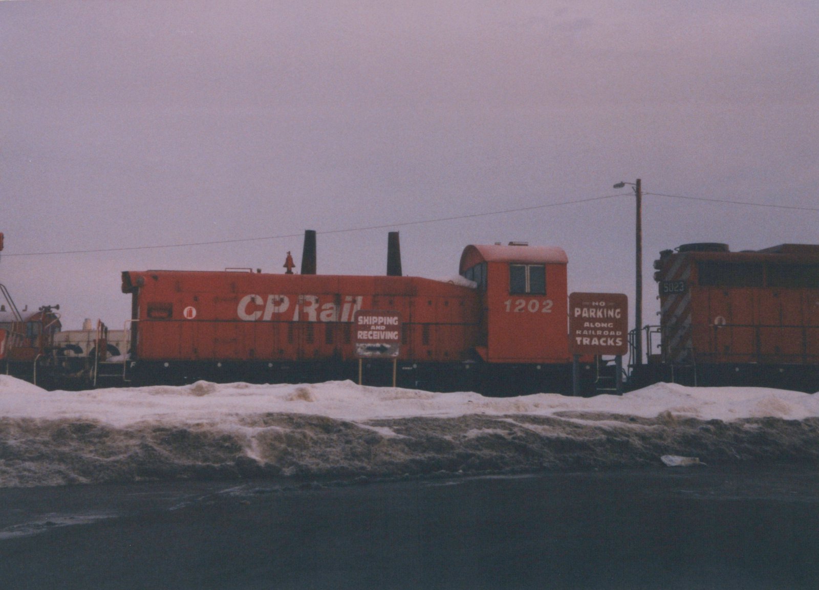 A side view of two red Canadian Pacific diesel locomotives, numbers 1202 and 5023, parked on a track next to snow-covered ground and signs reading 'Shipping and Receiving' and 'No Parking Along Railroad Tracks'.