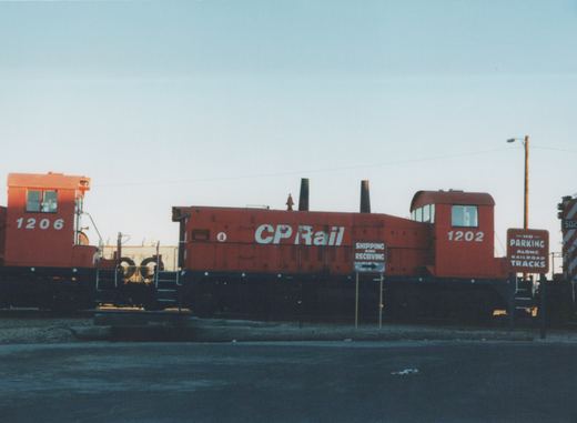 Two red Canadian Pacific locomotives, numbered 1206 and 1202, are parked on railroad tracks next to signs that read 'SHIPPING AND RECEIVING' and 'NO PARKING ALONG RAILROAD TRACKS'.