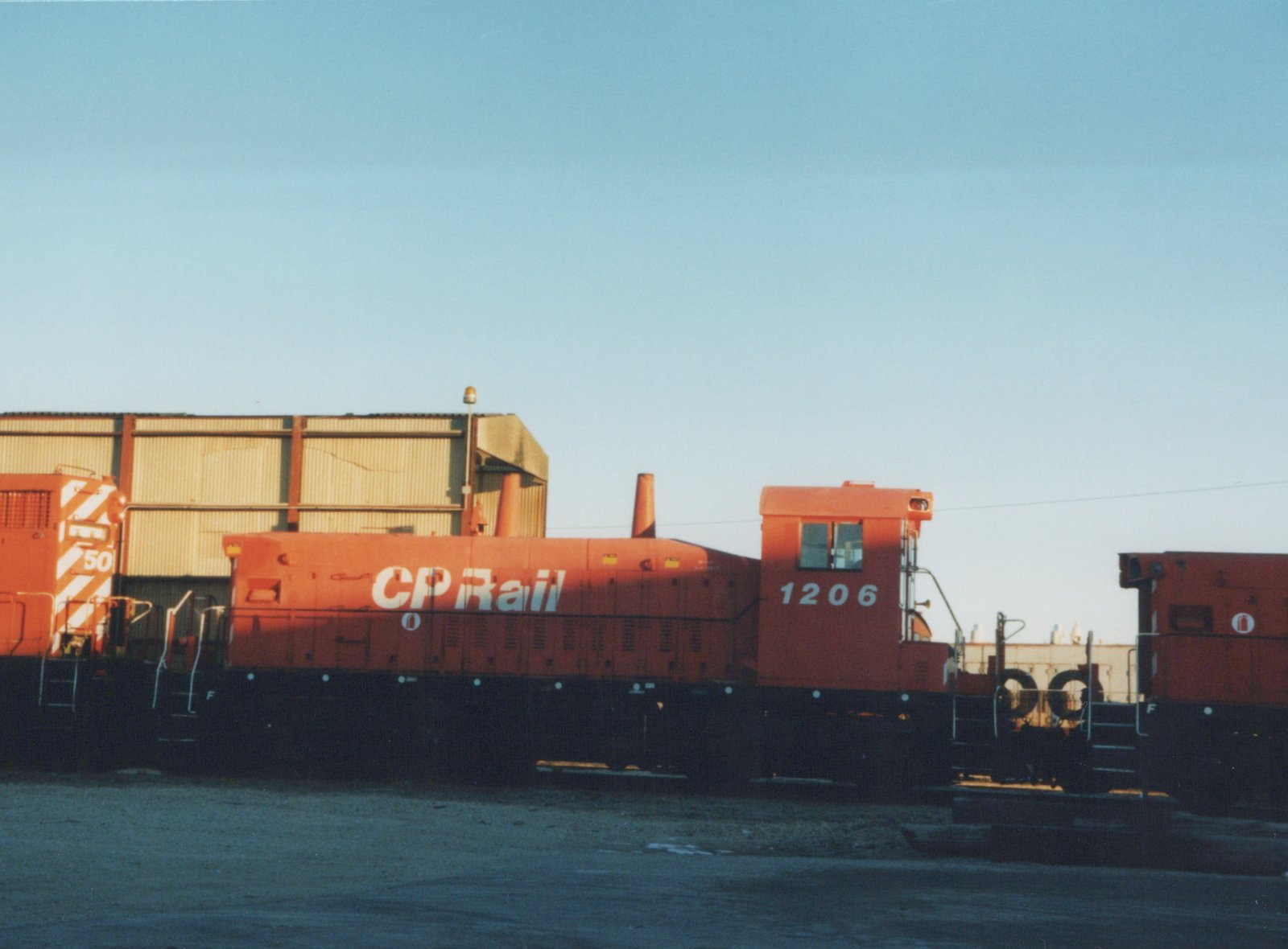 Side view of a Canadian Pacific locomotive number 1206 in orange paint, parked in a yard with another locomotive nearby under a clear blue sky.