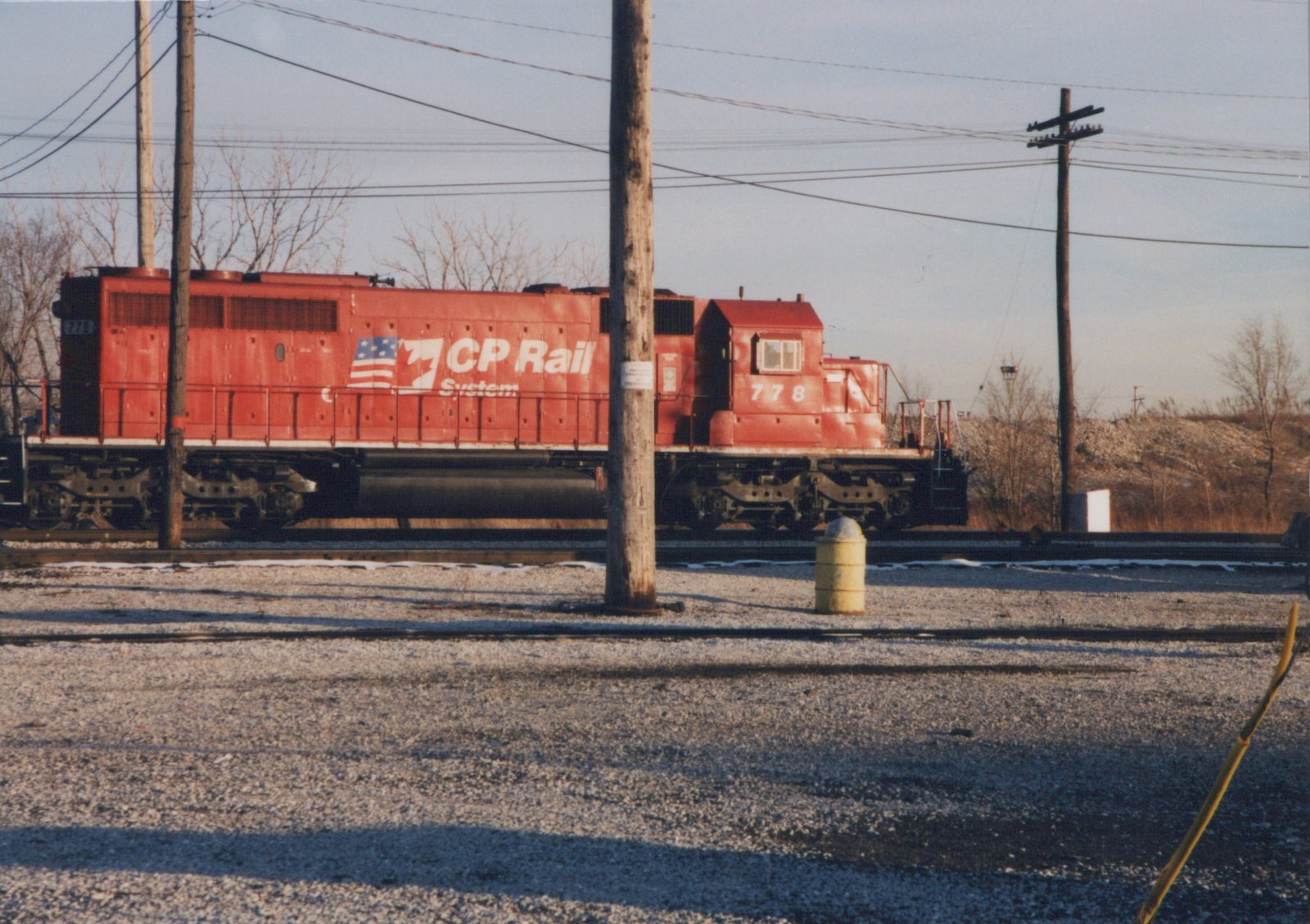 A red diesel locomotive with the number 778 and 'CP Rail System' lettering is parked in a gravel yard with bare trees and power poles, under a clear winter sky.