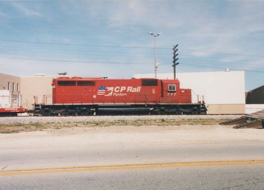 A red diesel locomotive with CP Rail System lettering and the number 777 sits on tracks beside a road, in front of an industrial building on a clear day.