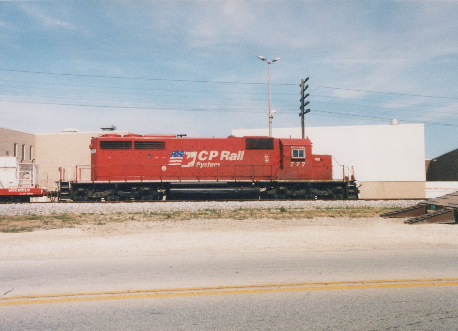 A red diesel locomotive with CP Rail System lettering and the number 777 sits on tracks beside a road, in front of an industrial building on a clear day.