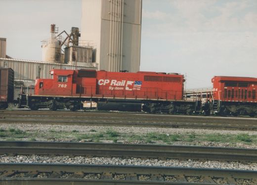 A red CP Rail System diesel locomotive numbered 762 is shown on a set of railroad tracks next to industrial buildings, with a second unit numbered 64 following behind.