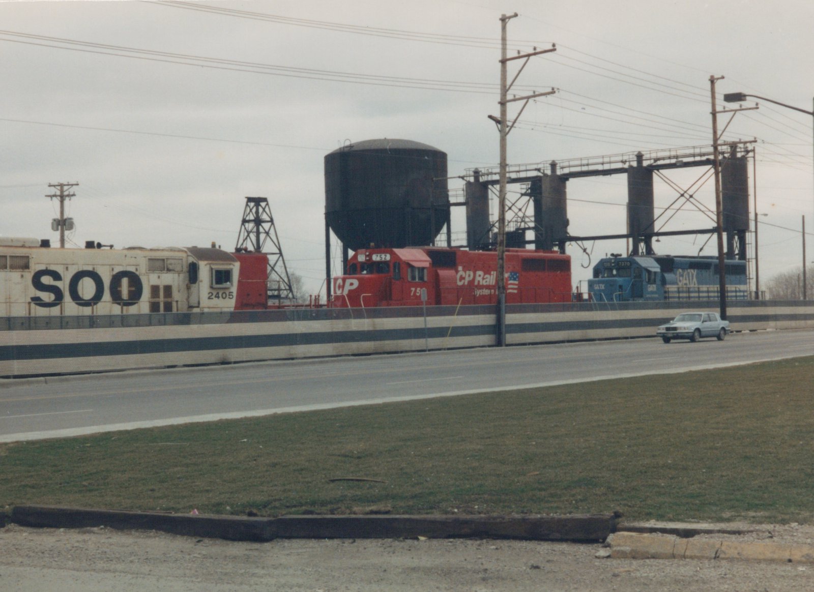 A scene showing three diesel locomotives—a white SOO Line 2405, a red Canadian Pacific 752, and a blue GATX 7370—on elevated tracks beside a road with a car driving by, under an overcast sky.
