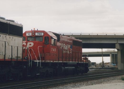 A red CP Rail System locomotive numbered 740 is pictured on tracks under a highway overpass on an overcast day, with construction visible in the background.