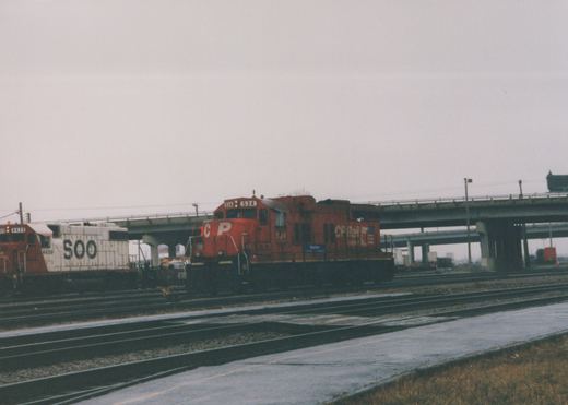Two diesel locomotives, one red CP Rail System 534 and one white Soo Line 4439, are parked in a railroad yard next to an elevated highway on a cloudy winter day.
