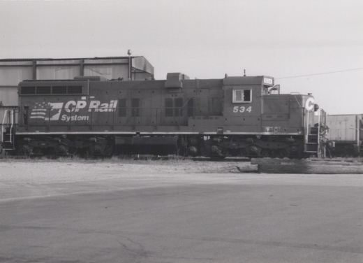 Side view of a CP Rail System diesel locomotive numbered 534 in a rail yard with industrial buildings in the background.